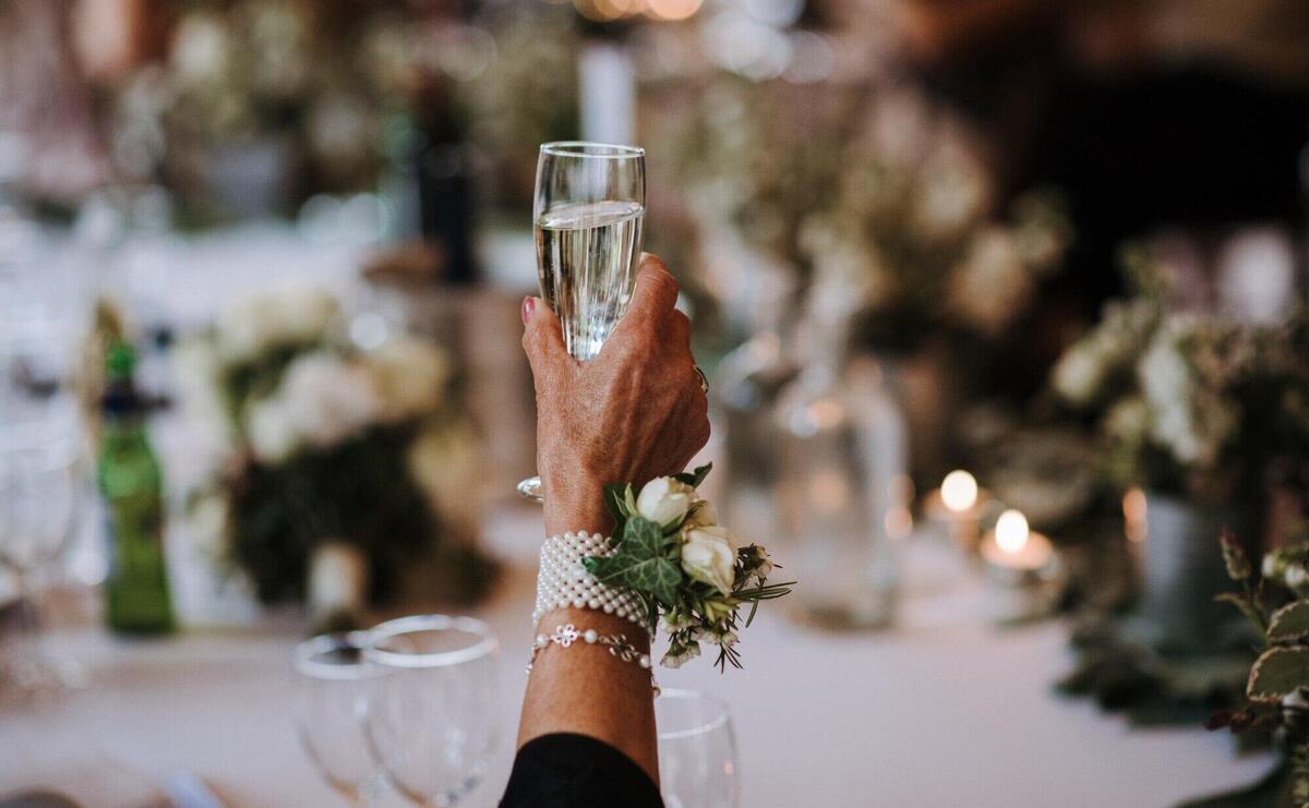 An old female holding a glass of champagne with a flower pinned on an accessory on her hand