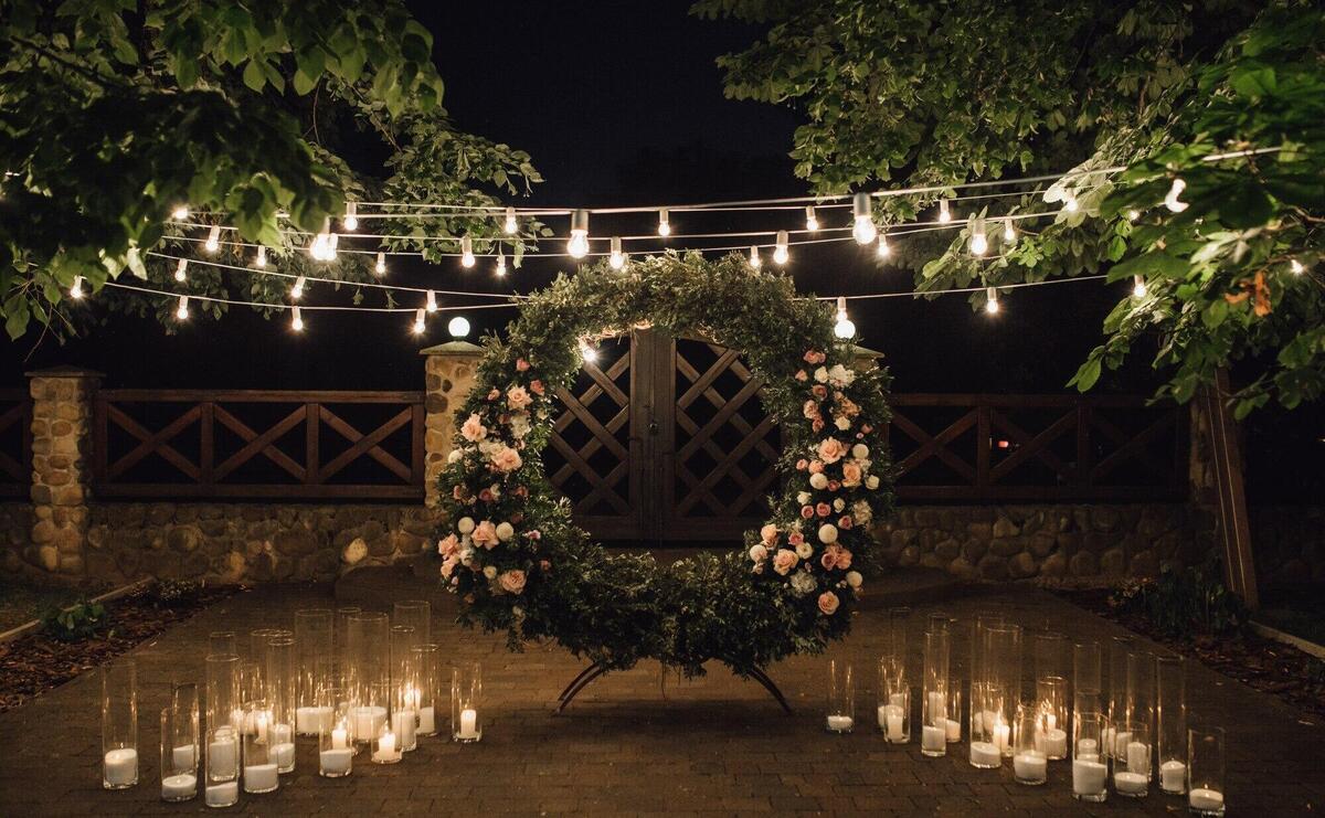 Beautiful photozone with big wreath decorated with greenery and roses in centerpiece, candles on the sides, and garland hanged between trees