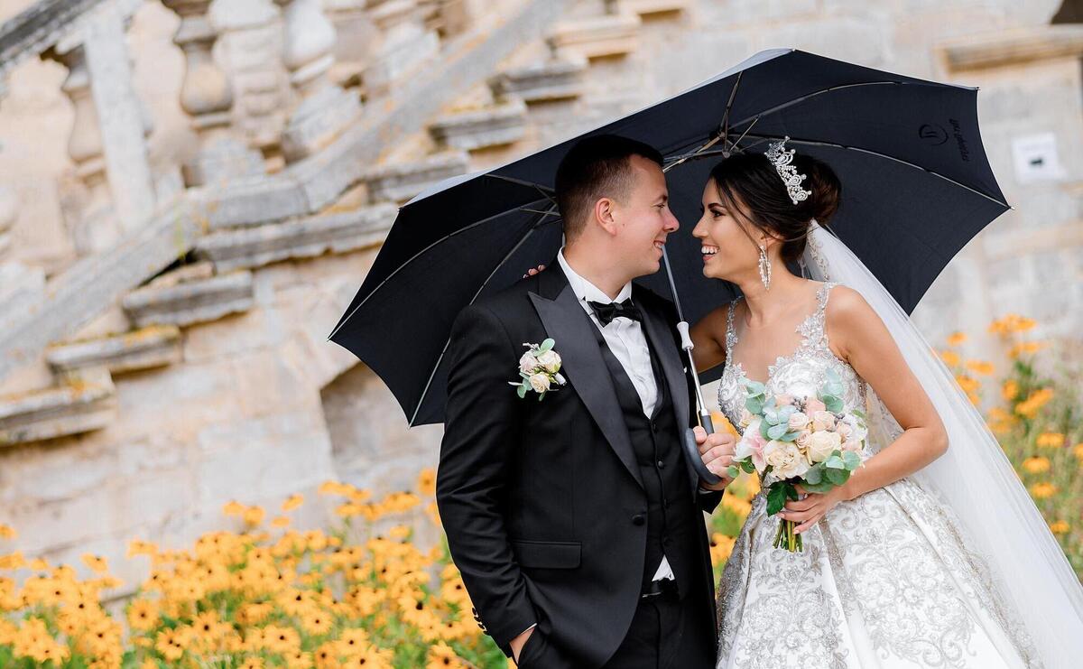 Front view of married man and woman smiling and looking to each other while standing under umbrella in rain weather on background of flowerbed and stairs of ancient building